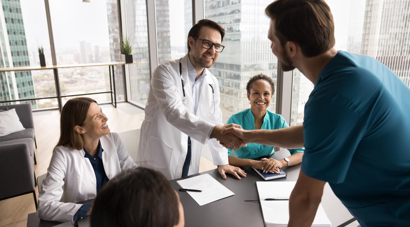 Smiling male doctor multiethnic medical group leader handshake with confident physician in blue surgeon uniform on staff briefing feel happy to meet young professional joining hospital