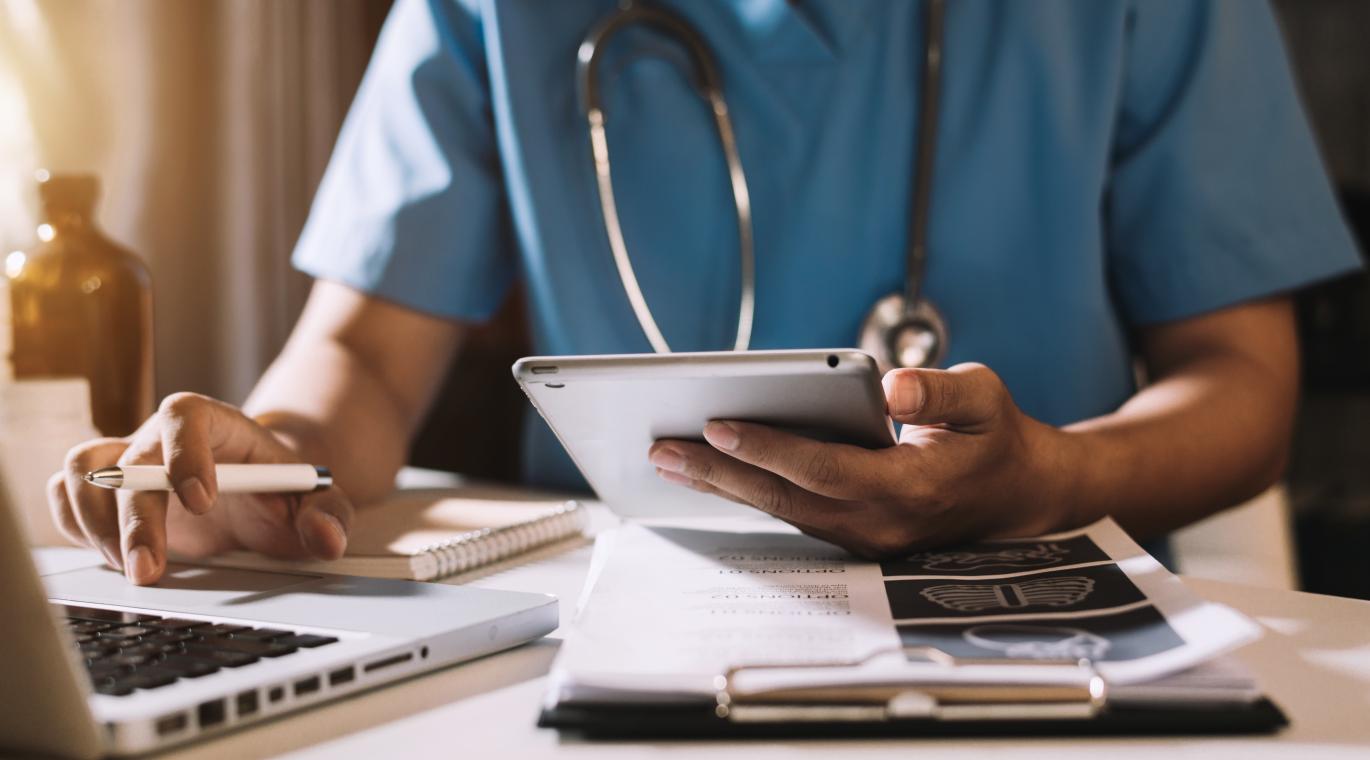 Physician working on a laptop and cellphone with papers on the desk