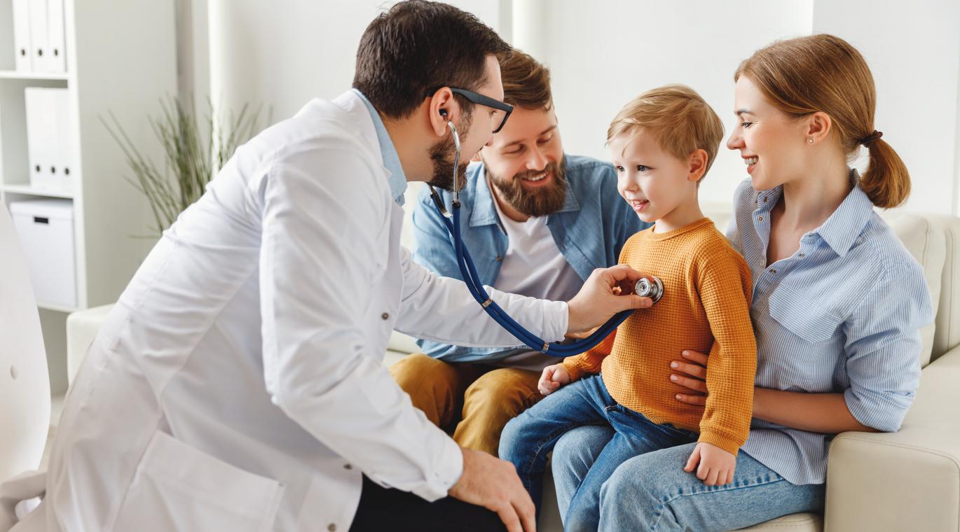 male physician using stethoscope on young child with family