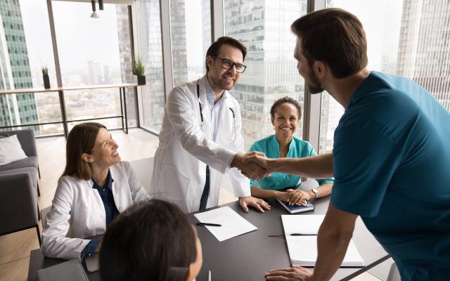 Smiling male doctor multiethnic medical group leader handshake with confident physician in blue surgeon uniform on staff briefing feel happy to meet young professional joining hospital