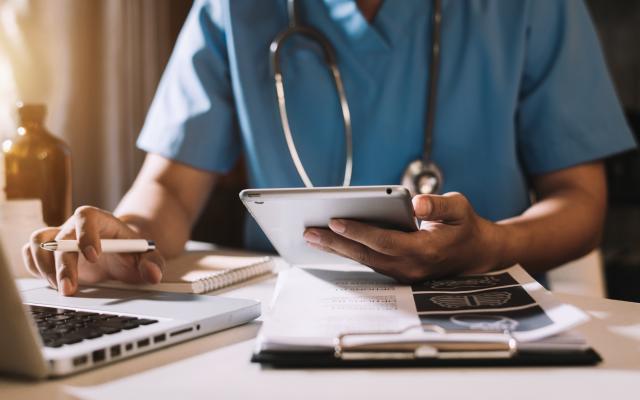 Physician working on a laptop and cellphone with papers on the desk