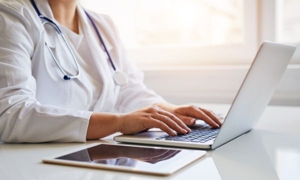 doctor typing on laptop with tablet next to them